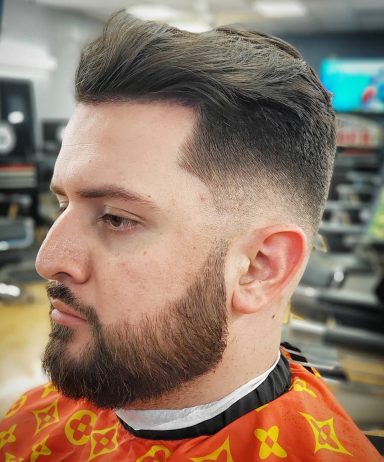 Profile shot of a man with a clean haircut and a well-groomed beard, wearing a barber's cape.