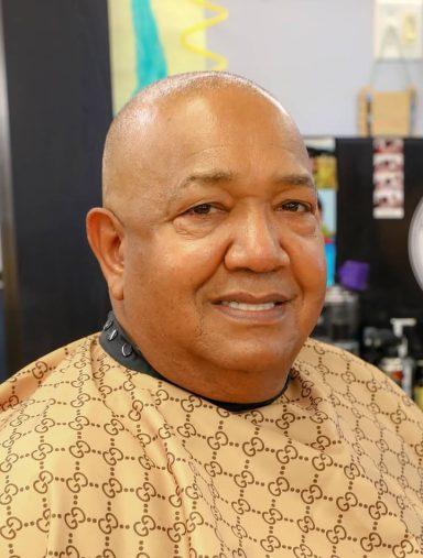 Smiling man with short hair in a barber shop, wearing a patterned cape.