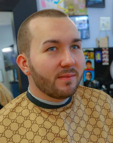 Young man with a short beard and buzz cut, wearing a patterned cape in a barbershop setting.