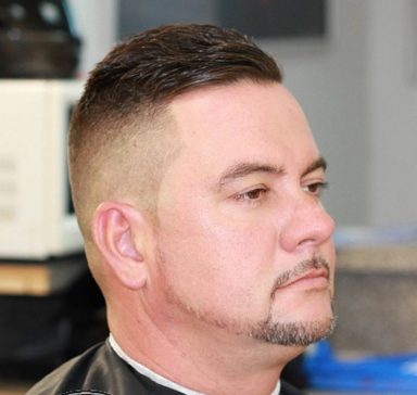 A man with a short haircut and facial hair, sitting in a barbershop.