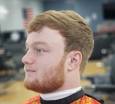 Young man with short, styled hair and a beard, seated in a barbershop.