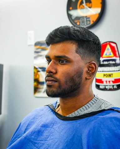 Man with short hair and beard sitting in a barber's chair, wearing a blue cape.