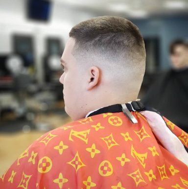 A young man with a fade haircut, wearing a patterned barber cape, sits in a salon.