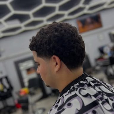 Young man with a curly haircut, sitting in a barbershop, wearing a patterned cape.