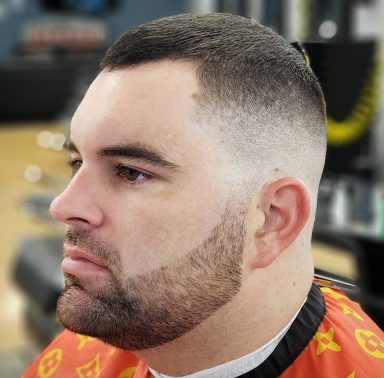 Man with a closely cropped fade haircut and a neatly trimmed beard, wearing a barber's cape.