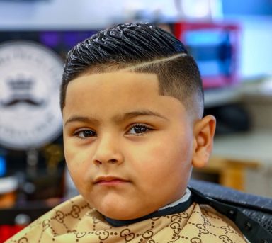 Young boy with a stylish haircut, wearing a barber's cape, looking at the camera.