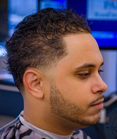 Man with curly hair and a neatly trimmed beard, sitting in a barbershop.
