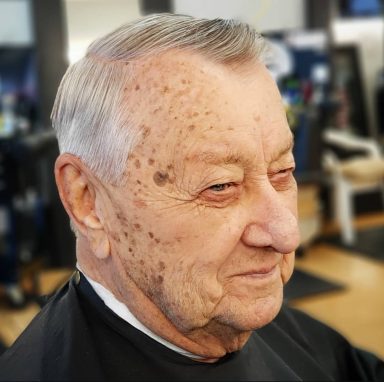 Elderly man with light gray hair and facial freckles, smiling in a barber shop setting.