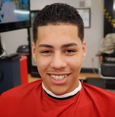 Smiling young man with short hair, wearing a red barber's cape in a barbershop setting.