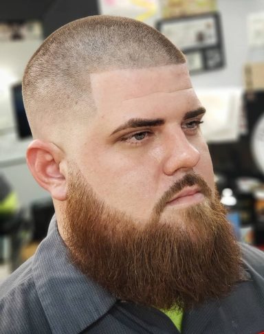 Close-up of a man with a shaved head and a thick beard, looking serious.
